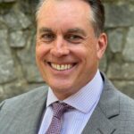 Smiling man in a suit and tie outdoors with stone wall background.