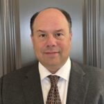 Professional man in suit and tie against a wood-paneled background.